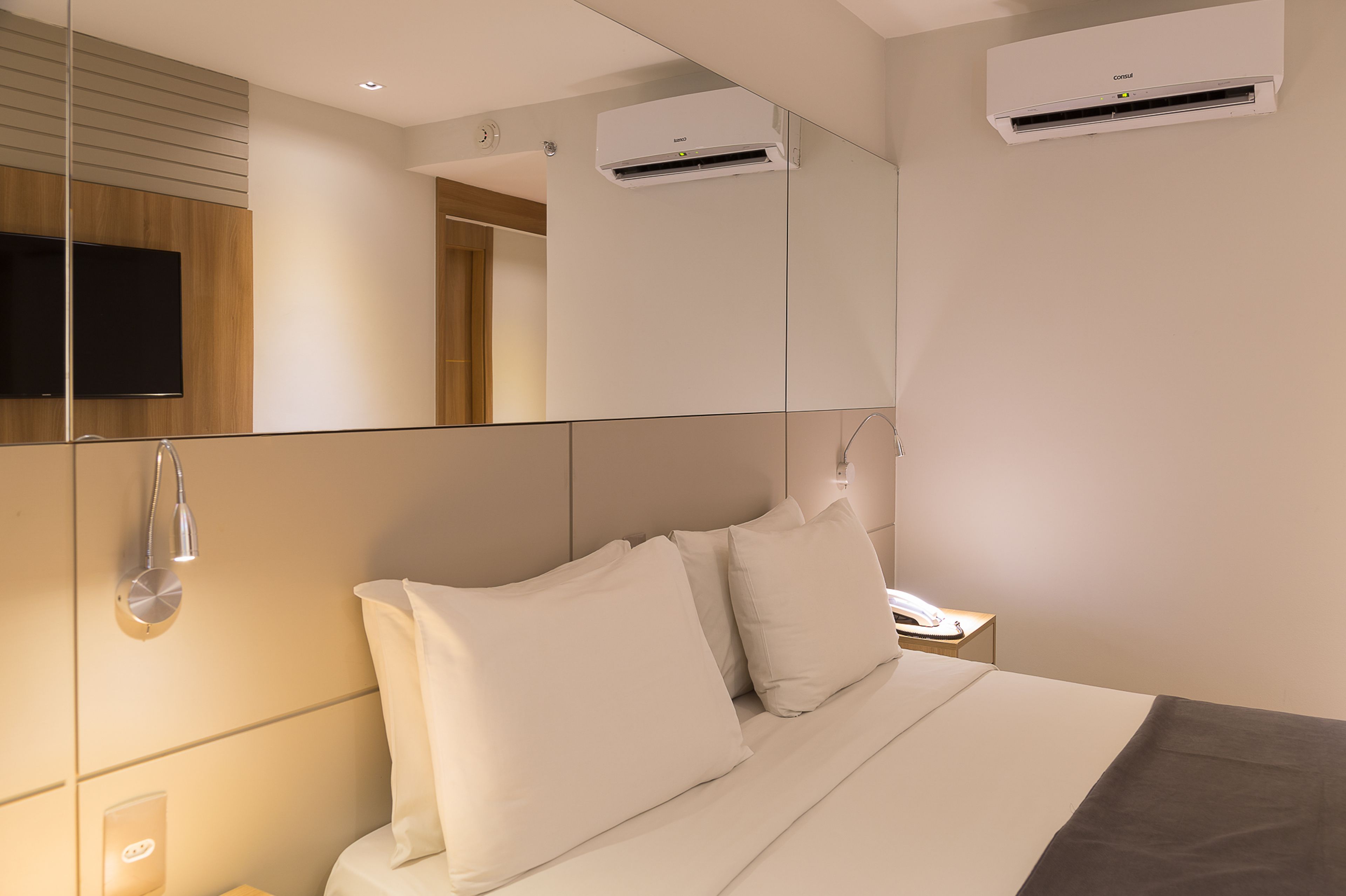 A modern hotel room with a large bed, a mounted TV, and a mirror reflecting the room.