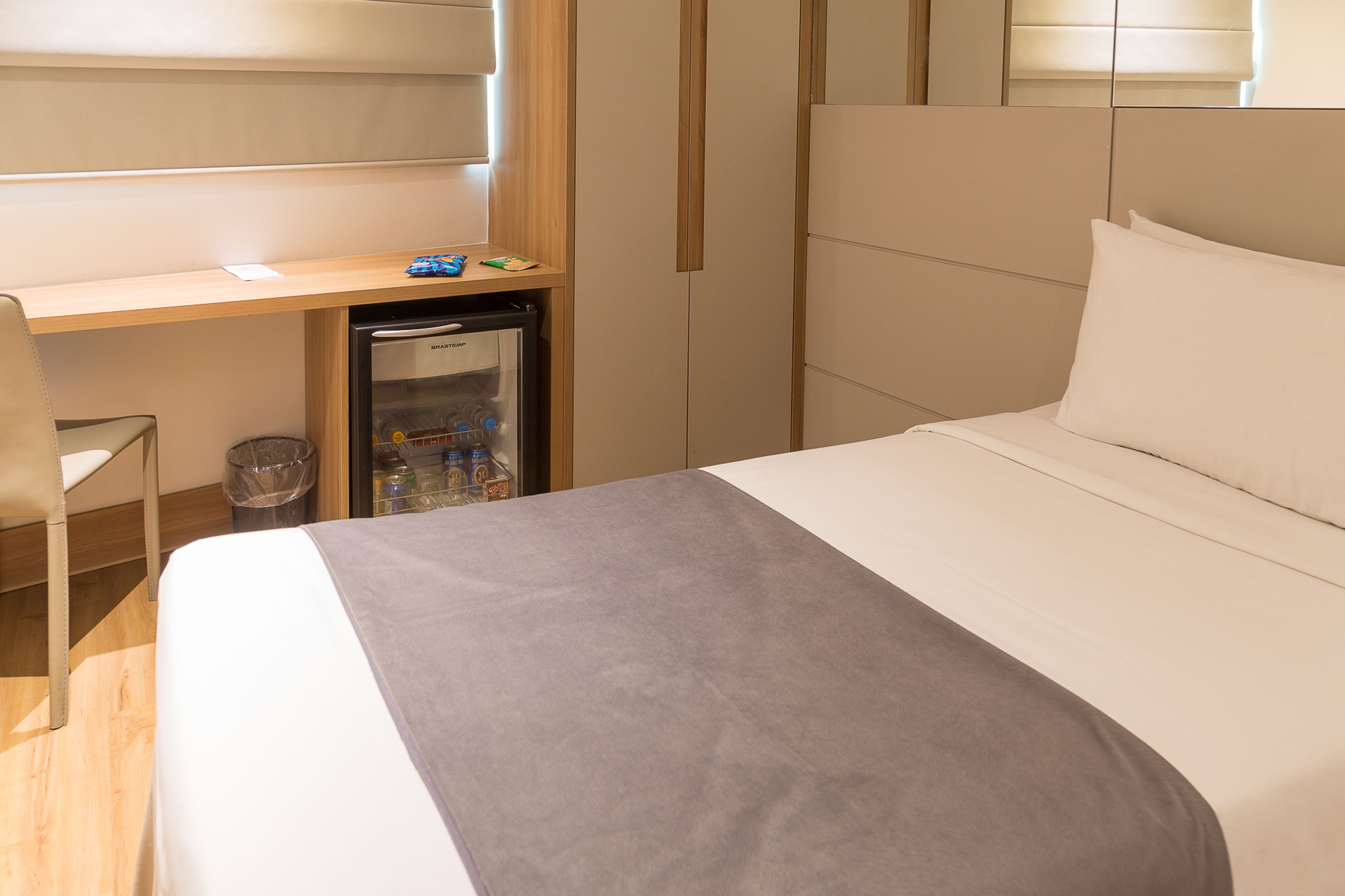 A hotel room with a bed, a desk, a small refrigerator, and a trash bin.