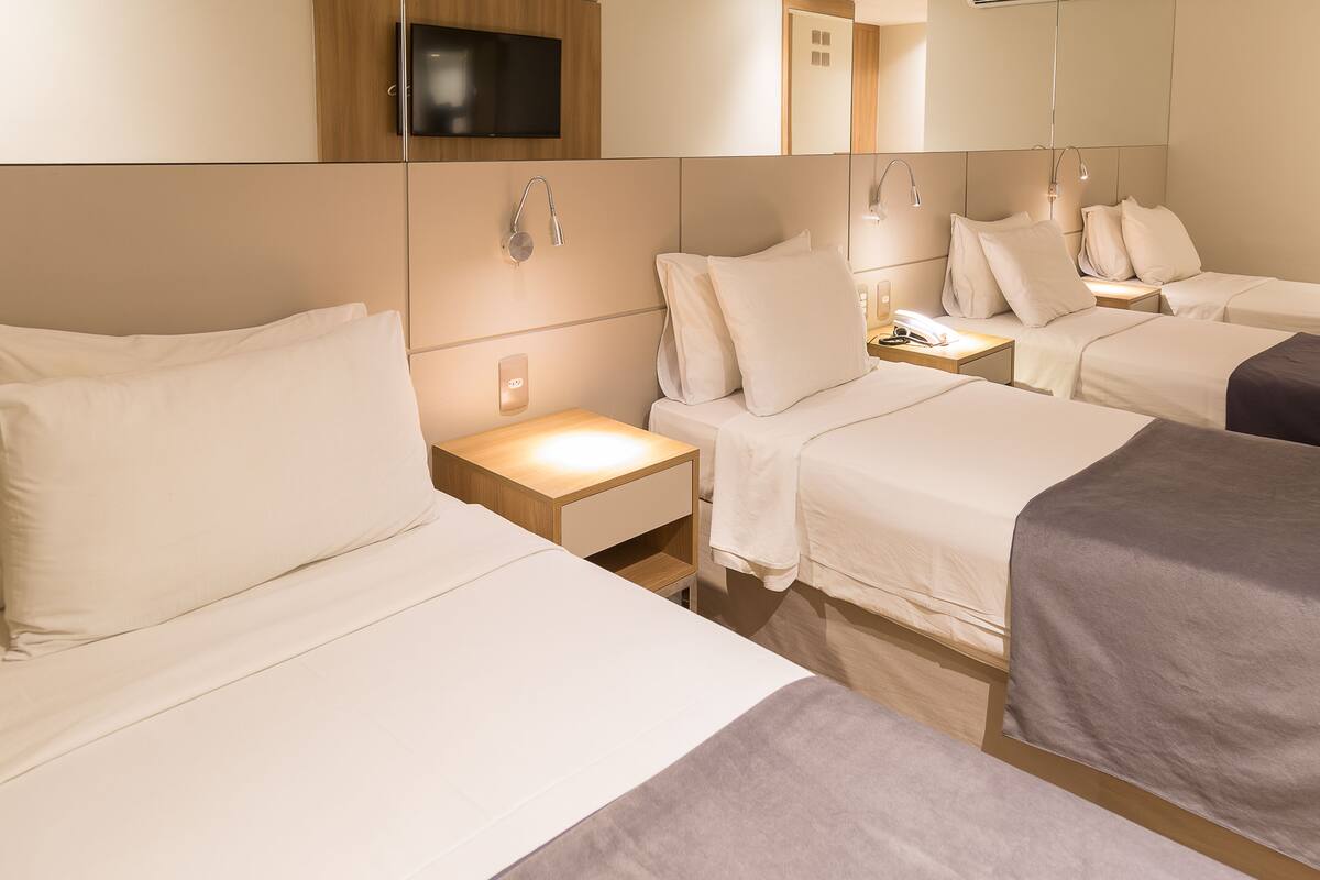 Habitación de hotel con tres camas, un televisor montado en la pared y una mesita de noche con lámpara.