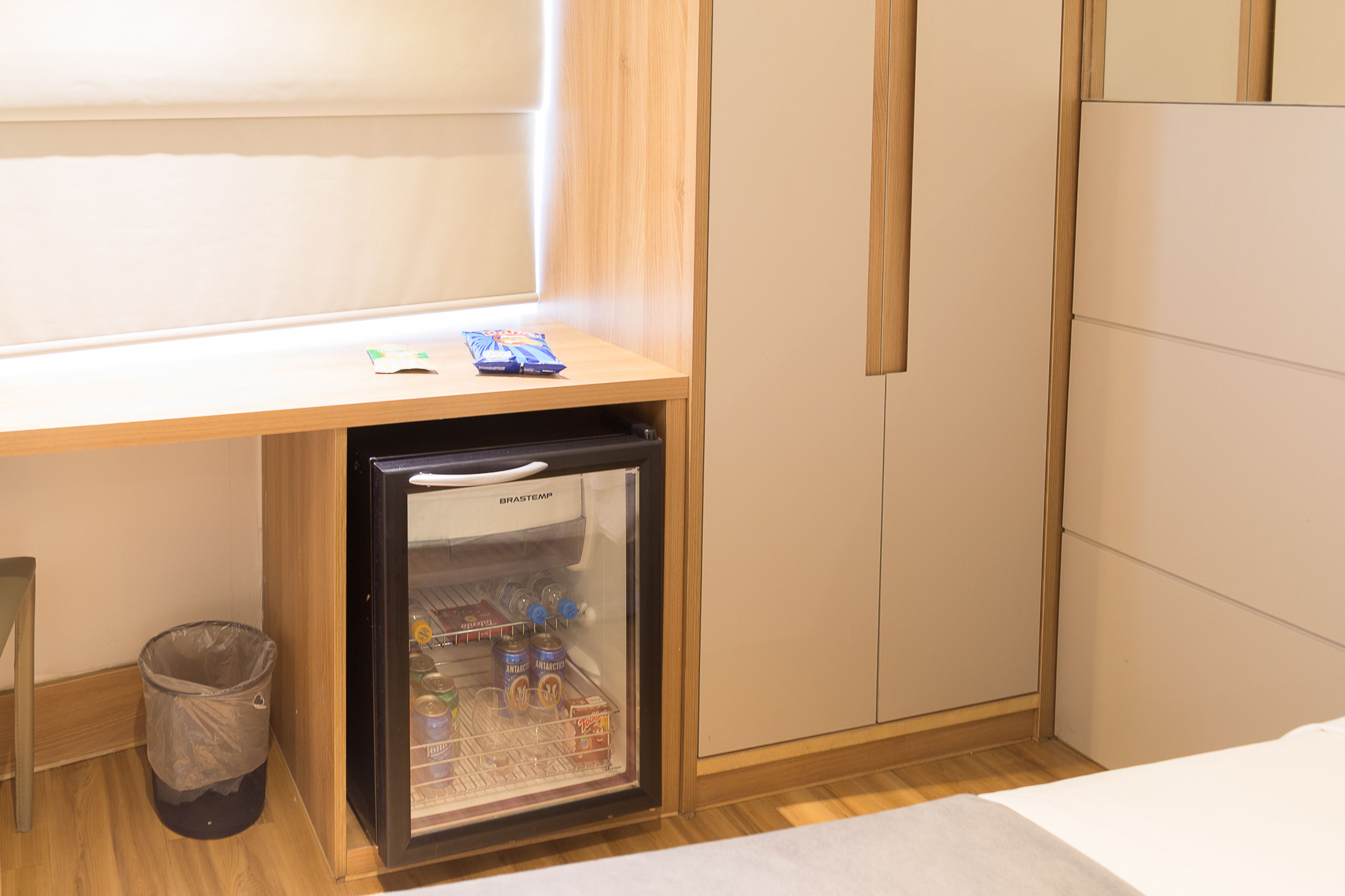 A compact refrigerator with beverages, a small trash bin, and a wooden desk with a window above.