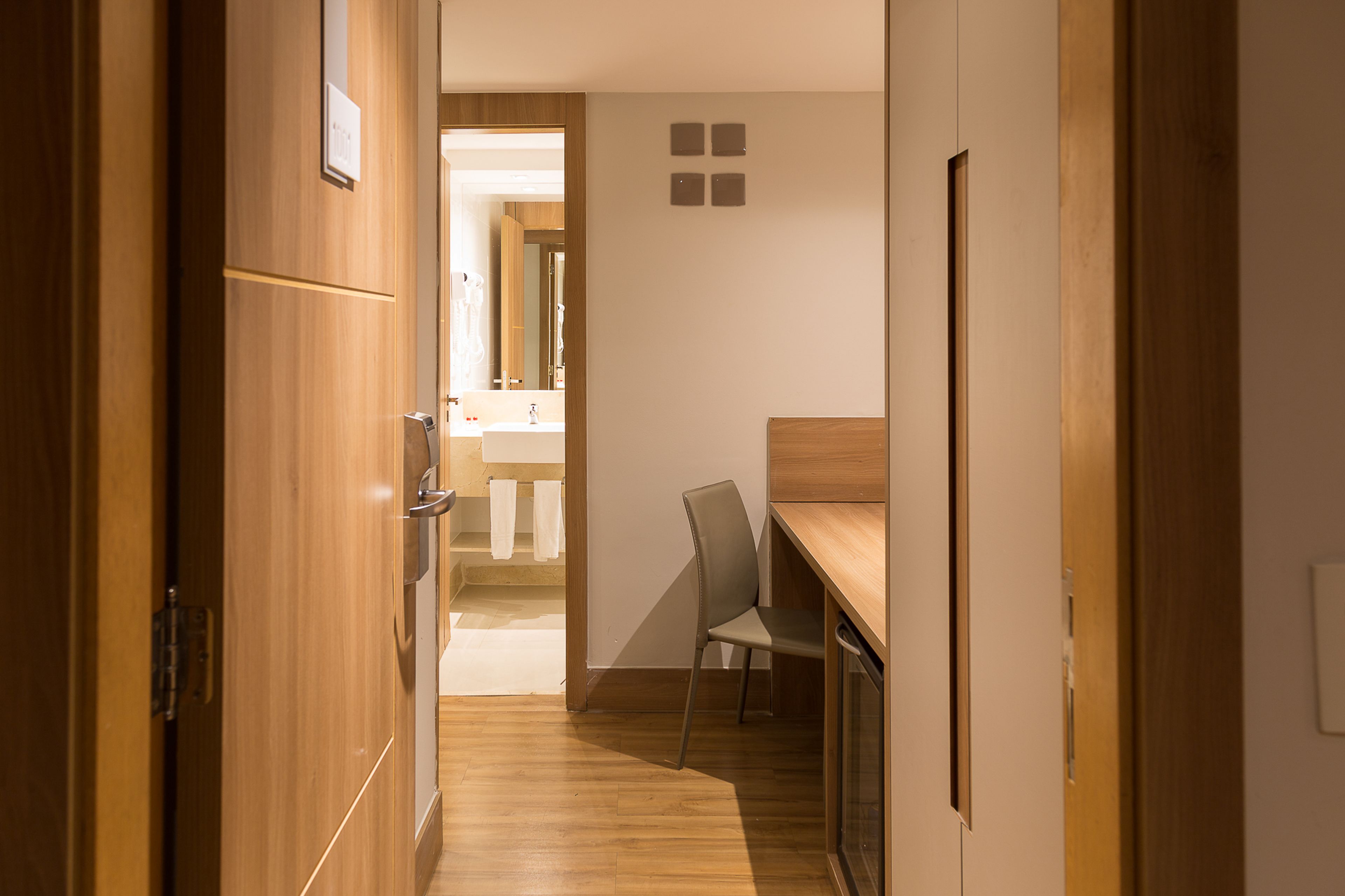 A hallway with wooden flooring, a desk with a chair, and a bathroom visible in the background.
