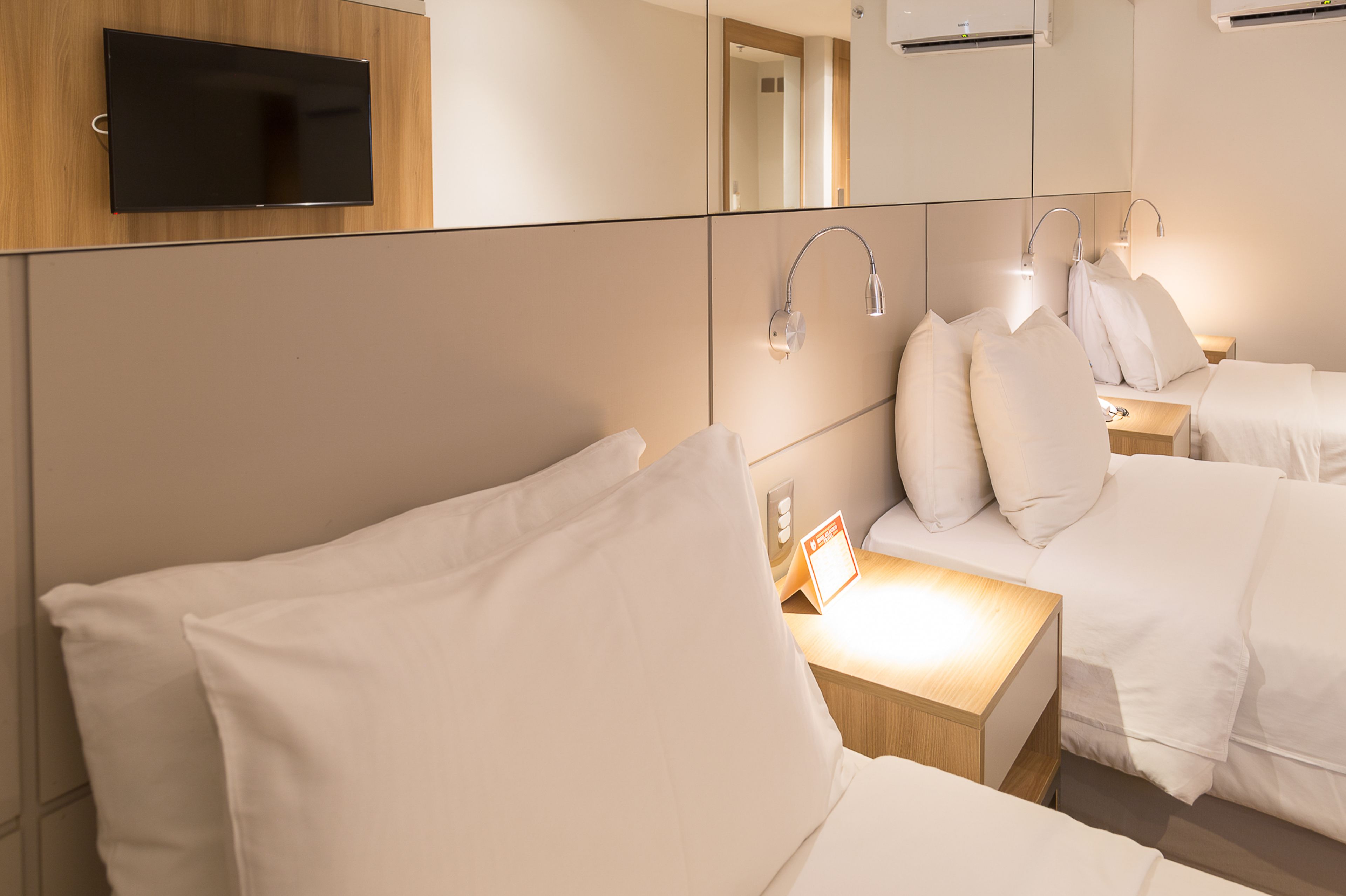 A hotel room with a TV mounted on the wall, a bedside table with a lamp, and two beds with white bedding.
