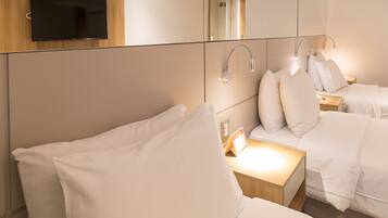 A hotel room with a TV mounted on the wall, a bedside table with a lamp, and two beds with white bedding.
