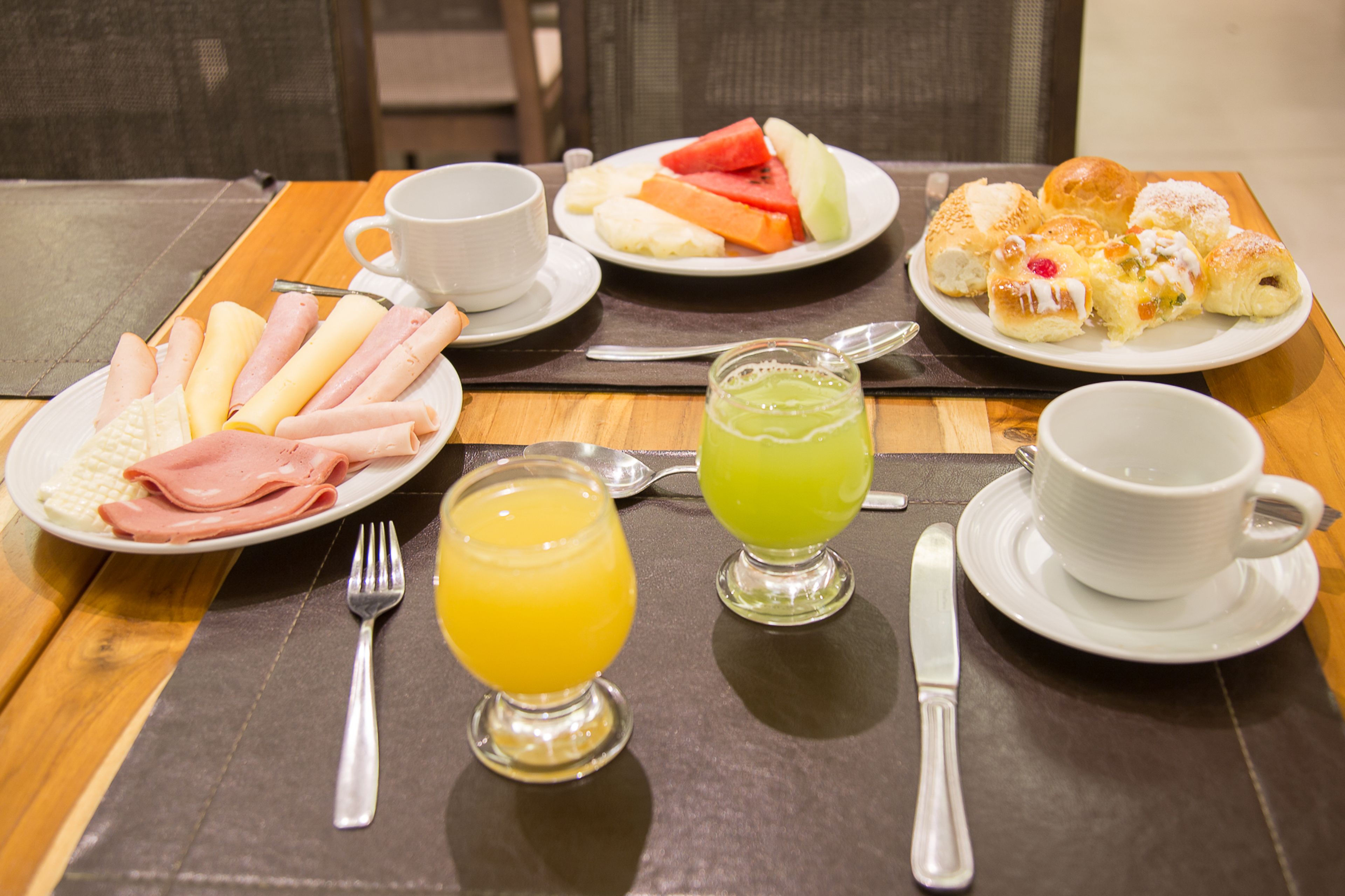 Una mesa de desayuno con platos de comida, una taza, un vaso de jugo de naranja y un vaso de jugo verde, además de cubiertos.