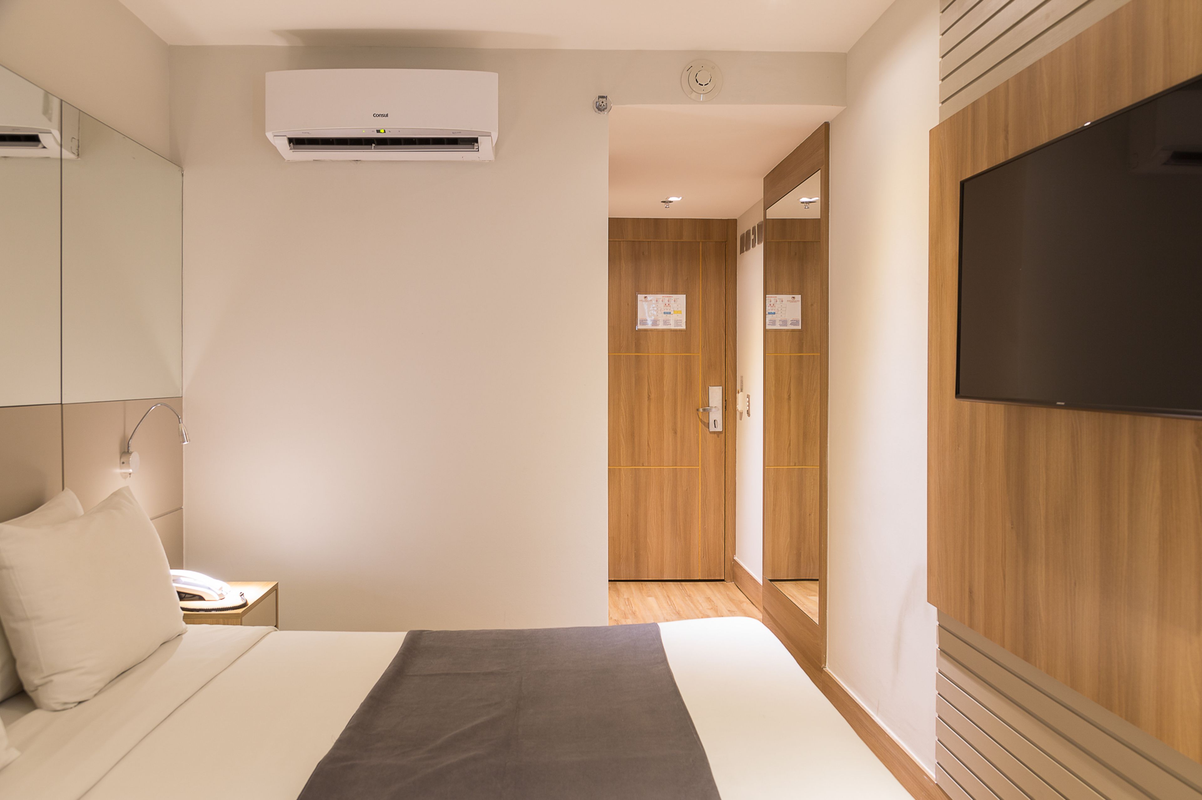 A hotel room with a bed, a bedside table with a lamp, a flat-screen TV mounted on the wall, and an air conditioning unit.