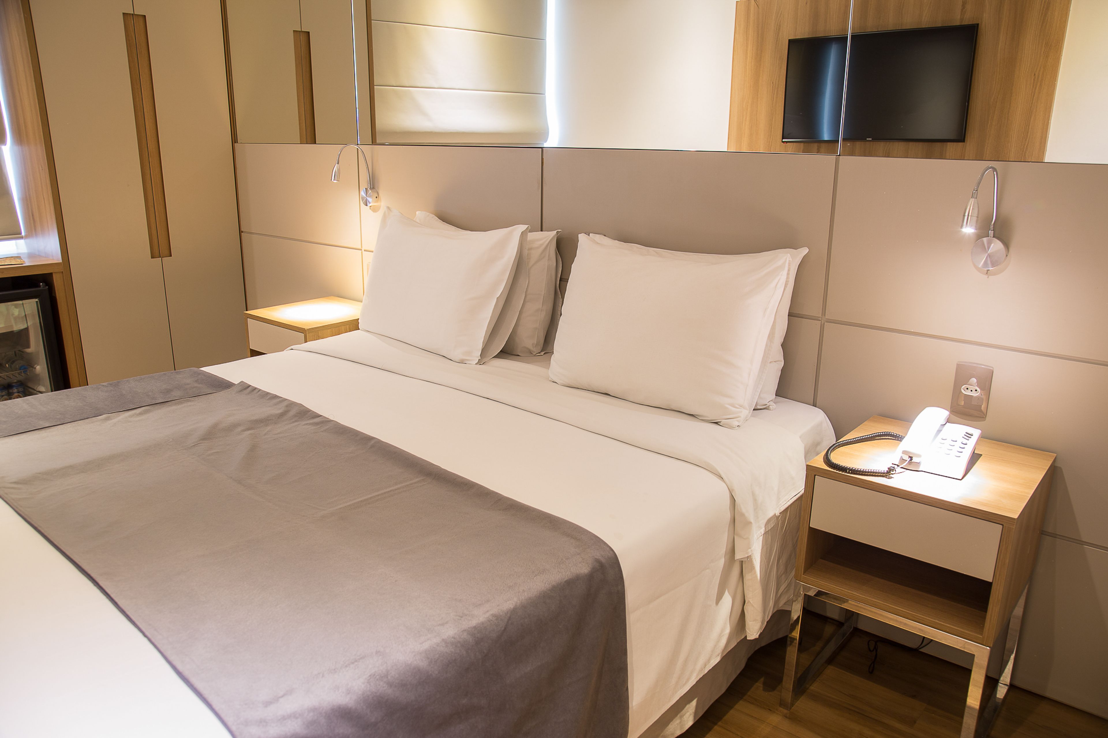 Una cama bien hecha con una colcha gris, una mesita de noche con una lámpara y un televisor montado sobre la cama.