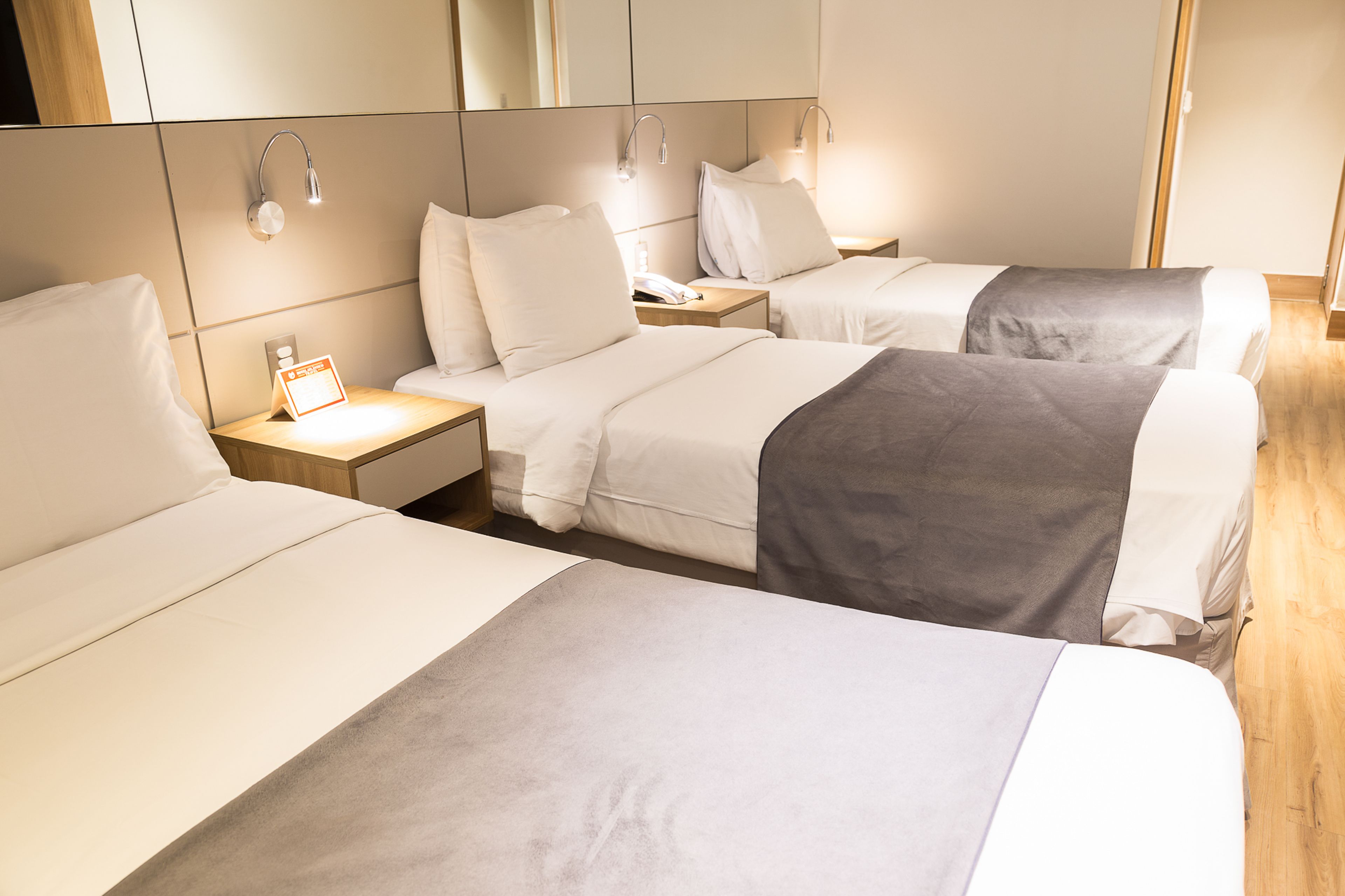 Een hotelkamer met drie bedden op rij, elk met een nachtkastje en een lamp.