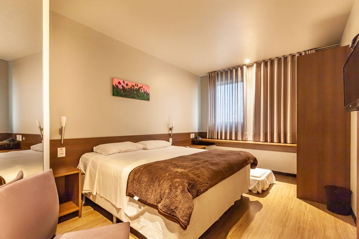 A hotel room with a large bed, bedside tables, a chair, a window with curtains, and a painting on the wall.