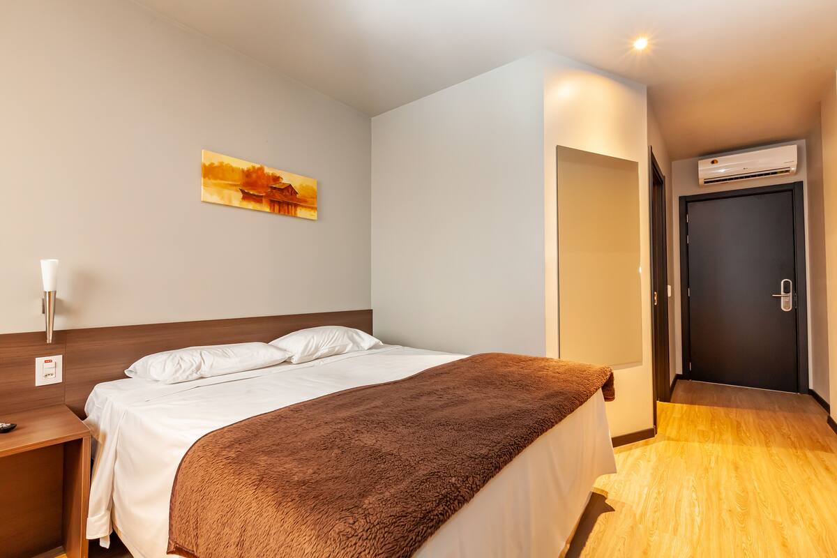 A hotel room with a bed, a nightstand, a painting on the wall, an air conditioning unit, and a door.