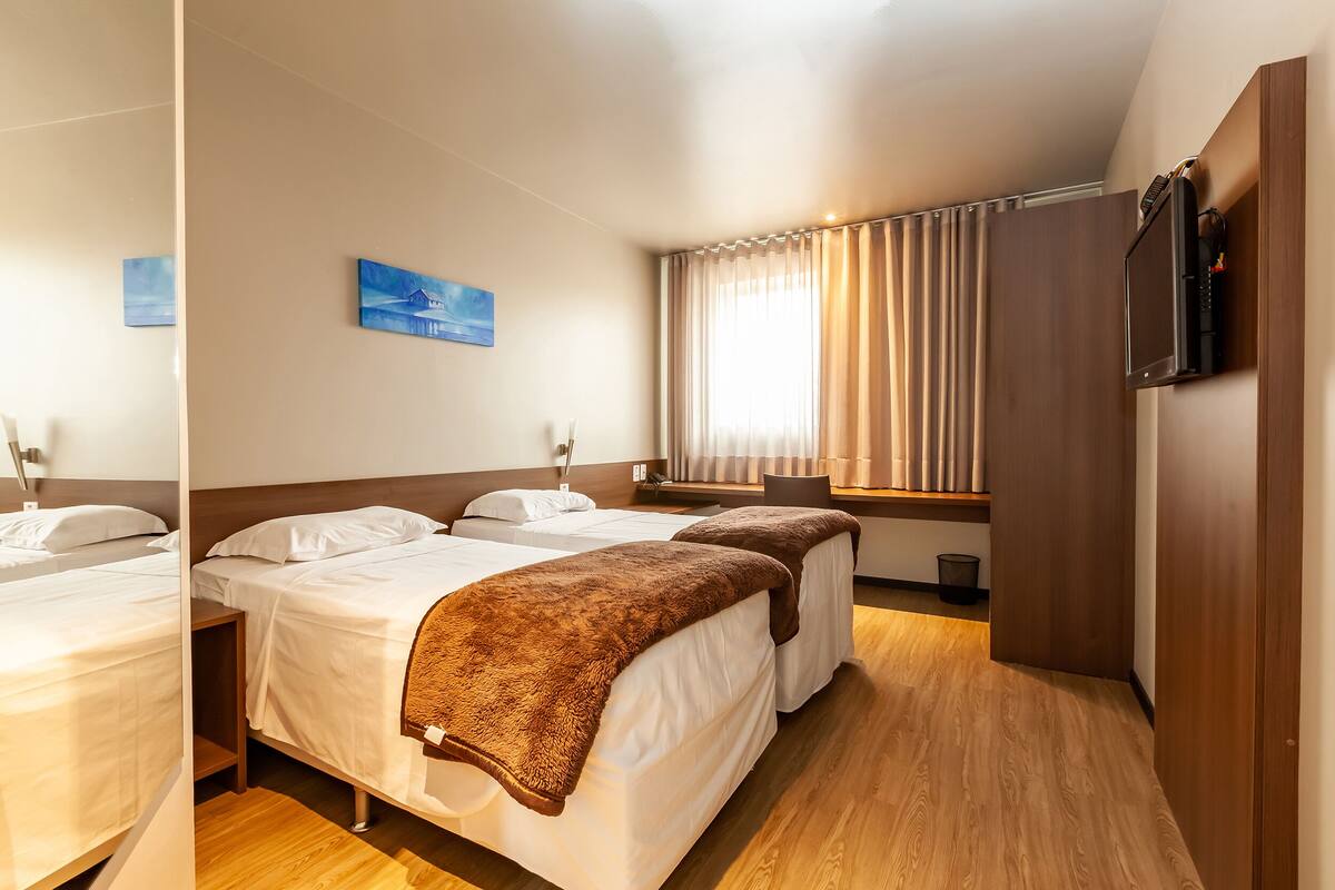 A hotel room with two beds, a TV mounted on the wall, a window with curtains, and a wooden floor.
