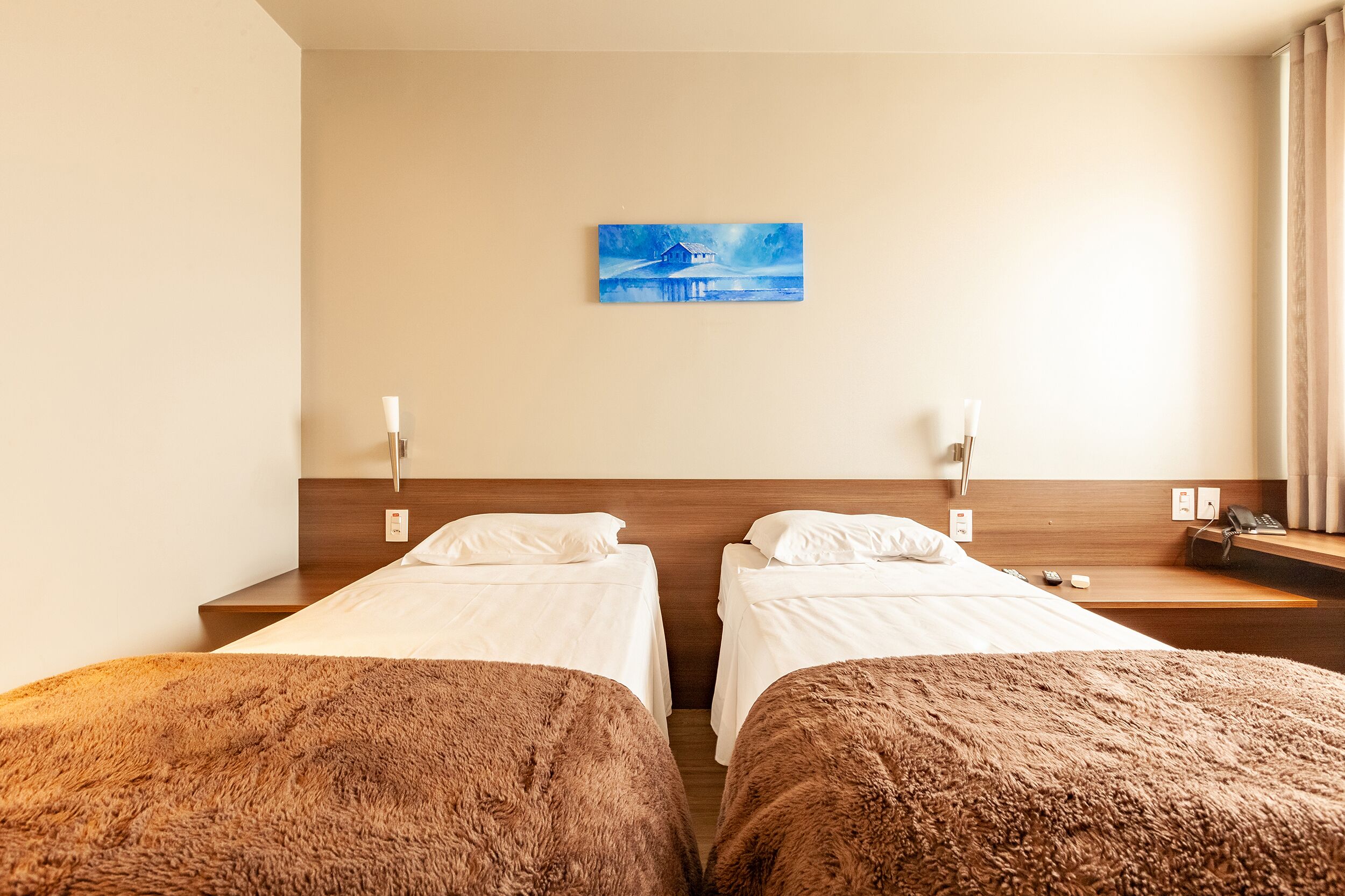 A hotel room with two single beds, a wooden headboard, a painting on the wall, and a bedside table with a lamp.