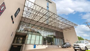 Un hotel moderno con fachada de vidrio, acceso cubierto y área de estacionamiento para automóviles visible.