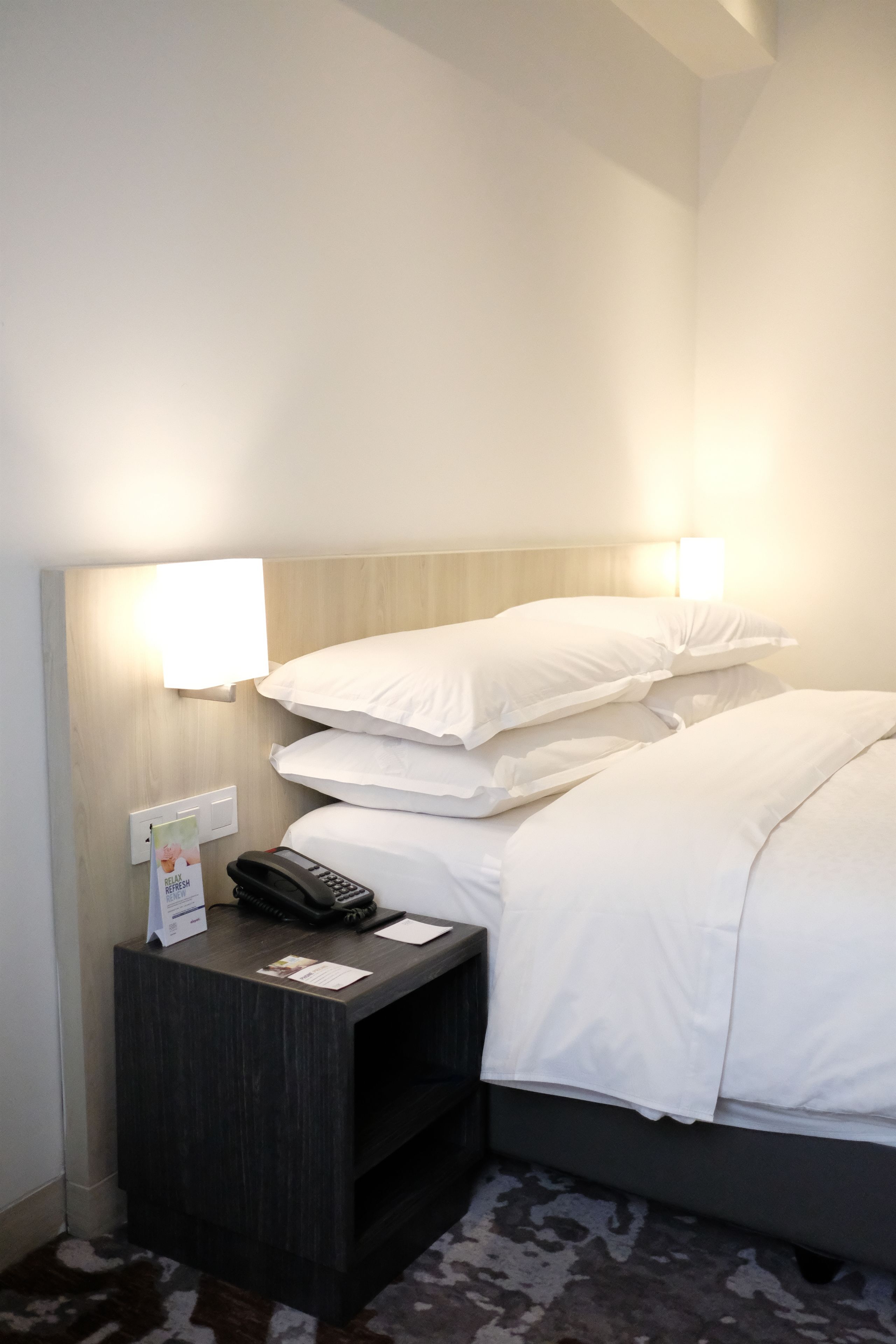 Ein Hotelzimmer mit Bett, weißen Kissen, einem Nachttisch mit Telefon und einer Wandleuchte.