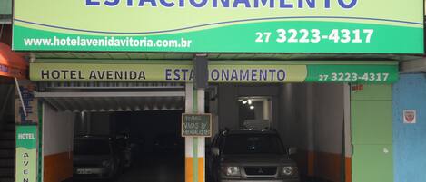 En hotelindgang med et skilt, der viser "HOTEL AVENIDA ESTACIONAMENTO" og en hjemmesideadresse "www.hotelavenidavistaoria.com.br".