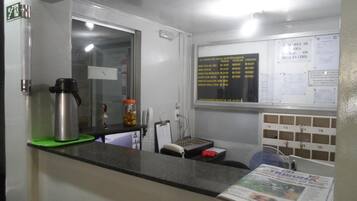 A hotel reception area with a counter, a coffee machine, a small refrigerator, a bulletin board with notices, and a newspaper.