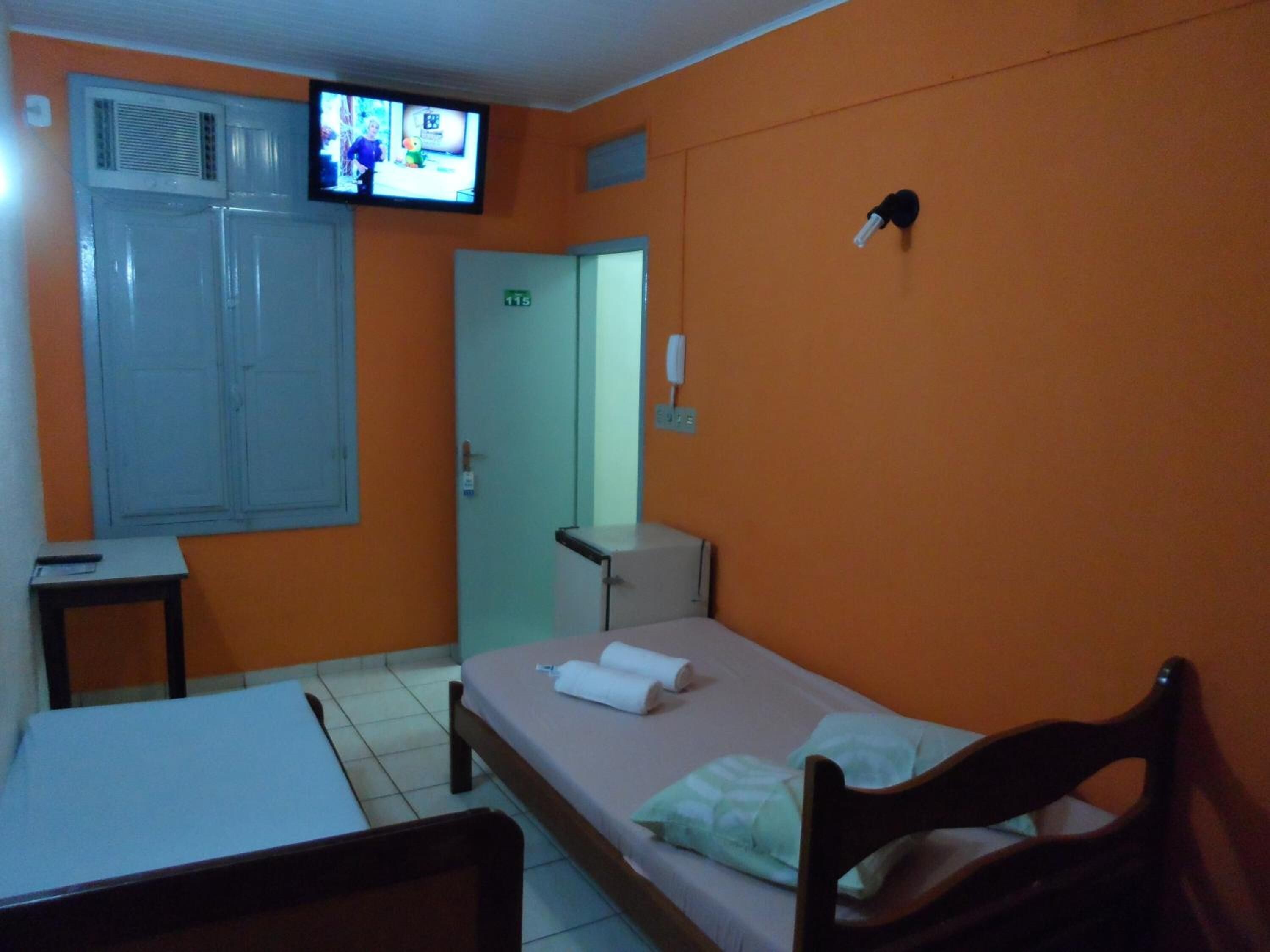 Quarto de hotel pequeno e individual, com uma cama, uma escrivaninha, televisão presa na parede e uma porta com placa indicando o número do quarto.