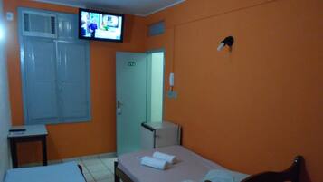 Quarto de hotel pequeno e individual, com uma cama, uma escrivaninha, televisão presa na parede e uma porta com placa indicando o número do quarto.