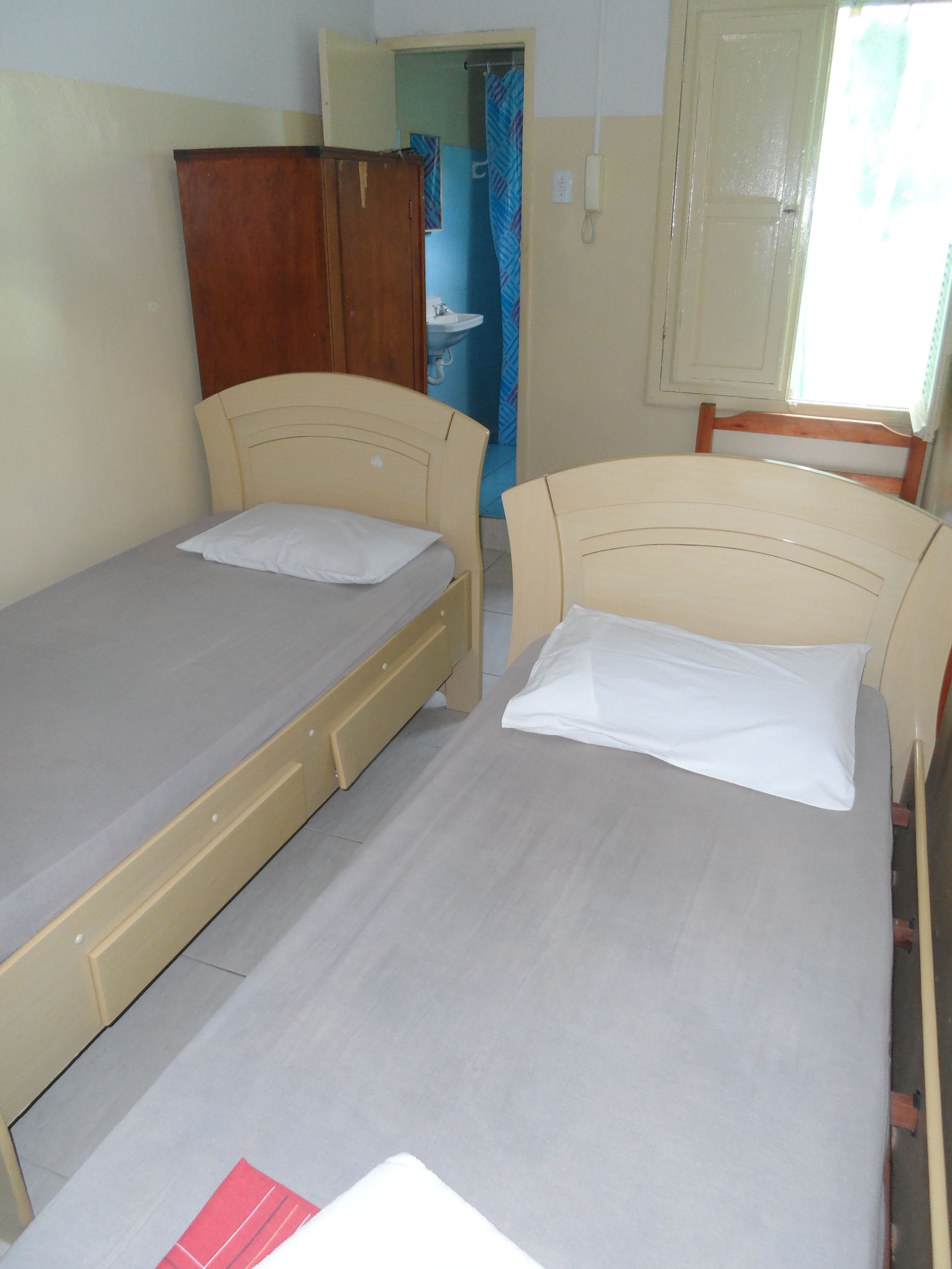 A room with two single beds, a wooden wardrobe, a bathroom visible through an open door, and a window with curtains.
