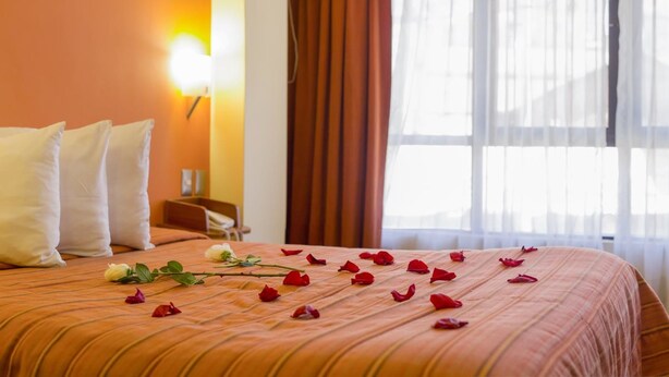 Una habitación de hotel con cama, almohadas, mesita de noche, lámpara y ventana con cortinas.