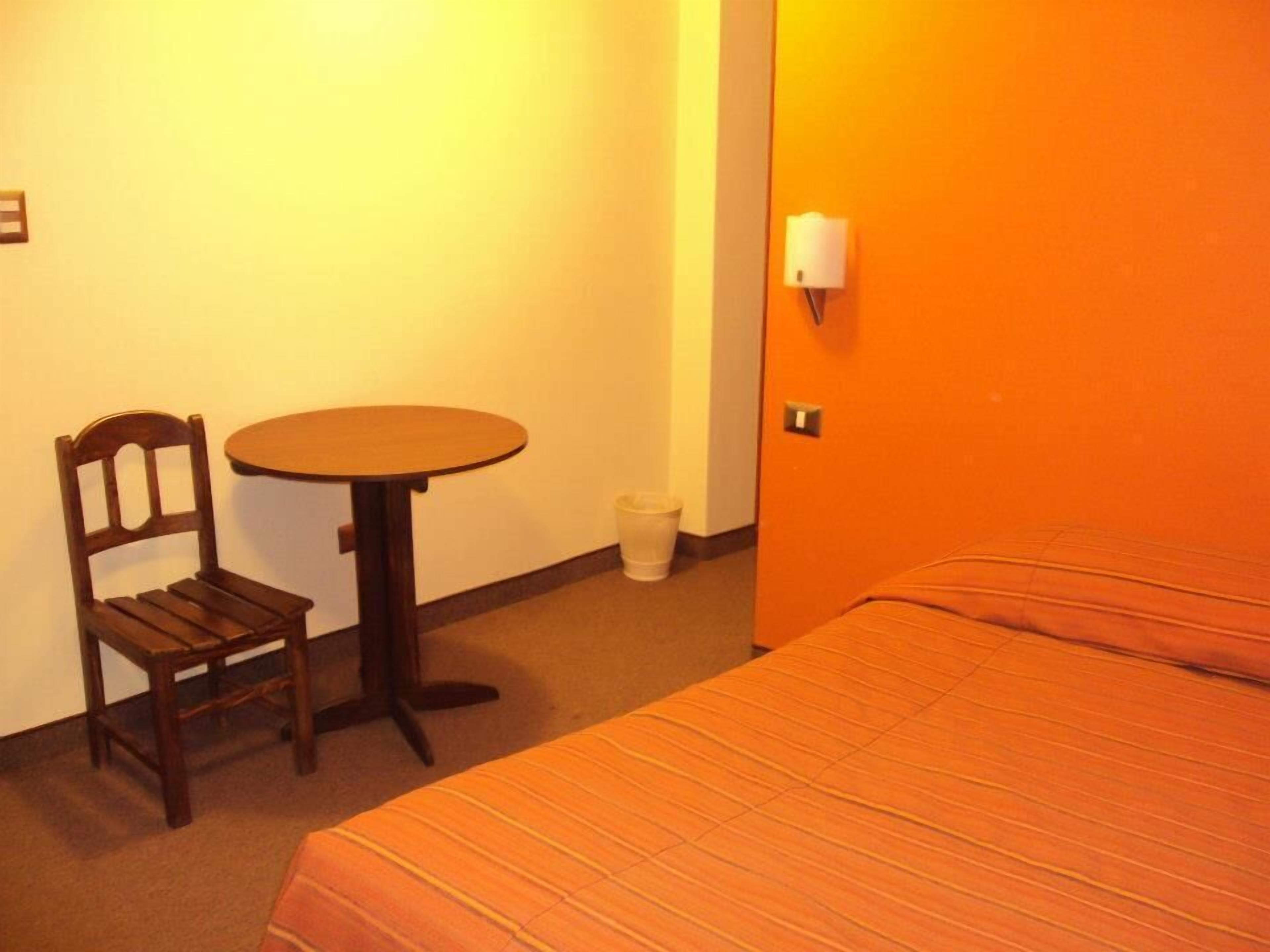 Una habitación de hotel con una cama, una silla de madera, una mesita redonda y un cubo de basura.