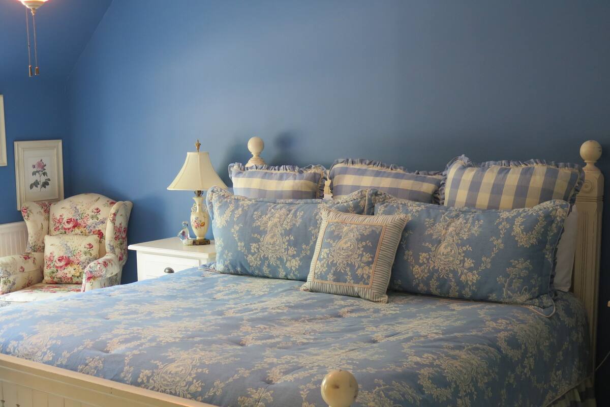Une chambre avec un mur bleu, un fauteuil à motifs floraux et un lit recouvert d’une literie bleue à motifs floraux.