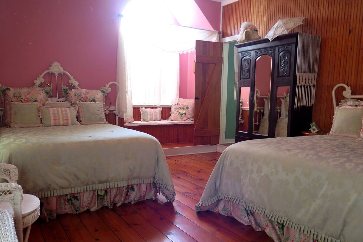 Une chambre avec deux lits, un plancher en bois, un grand miroir et une fenêtre avec des rideaux.