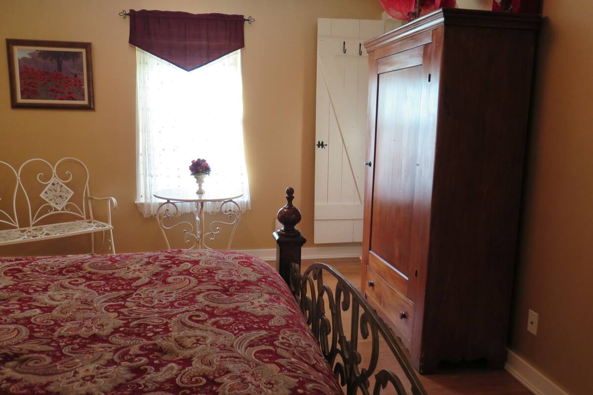 Une chambre à coucher avec un lit, une armoire en bois, une chaise en métal blanche, une petite table ronde avec un vase de fleurs et une fenêtre avec un rideau.