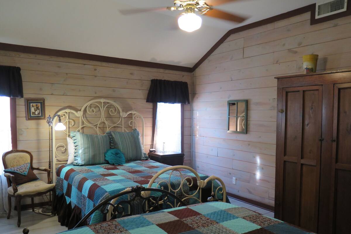 Une chambre avec un mur lambrissé en bois, un lit recouvert d’une courtepointe, une chaise, une armoire en bois et un ventilateur de plafond.