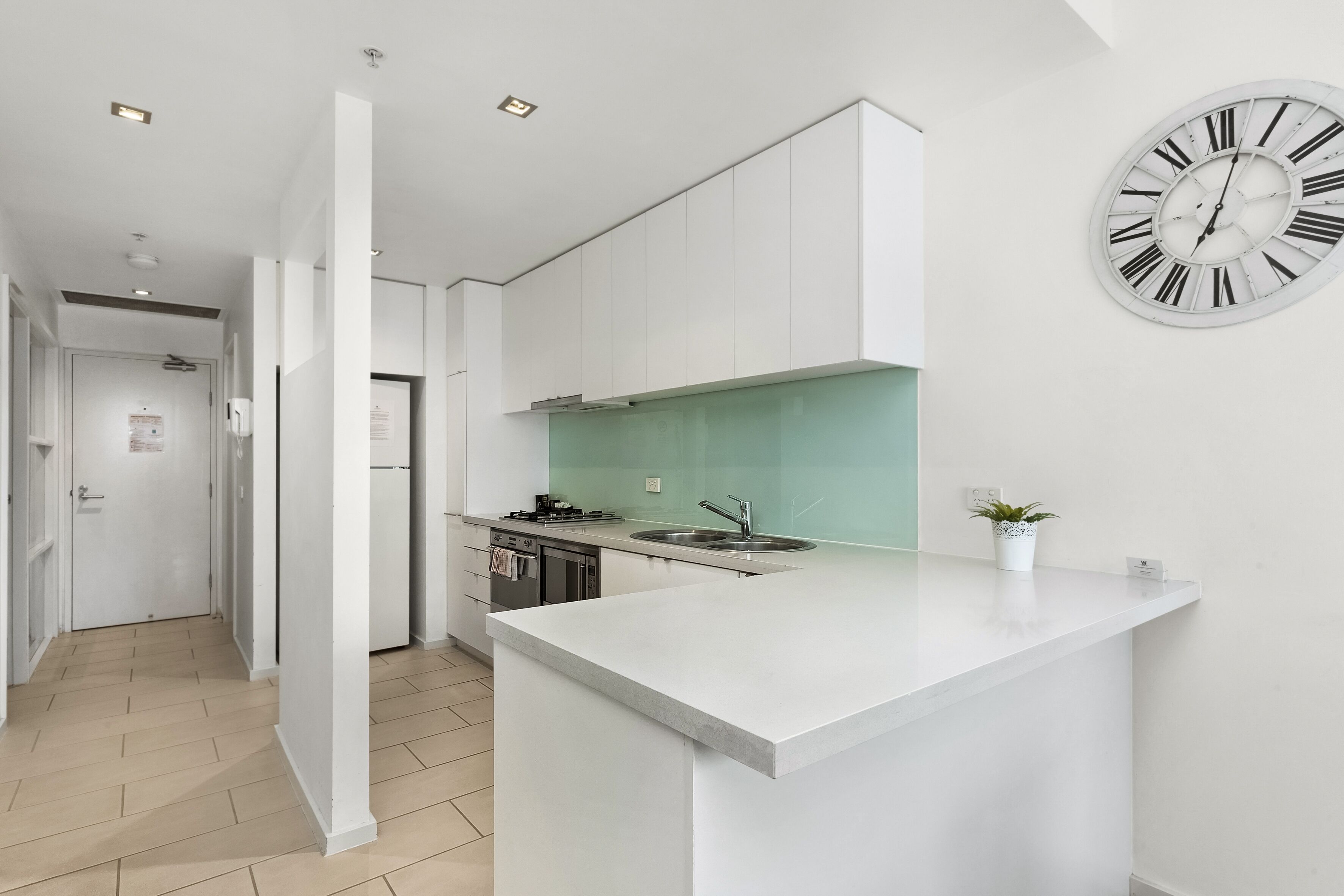 A modern kitchen with white cabinetry, a large island, a stainless steel sink, and a wall-mounted clock.