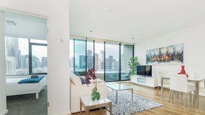 Ein modernes Wohnzimmer mit Sofa, Couchtisch und Fernseher, großen Fenstern mit Stadtblick sowie einem Essbereich mit Stühlen und Tisch.