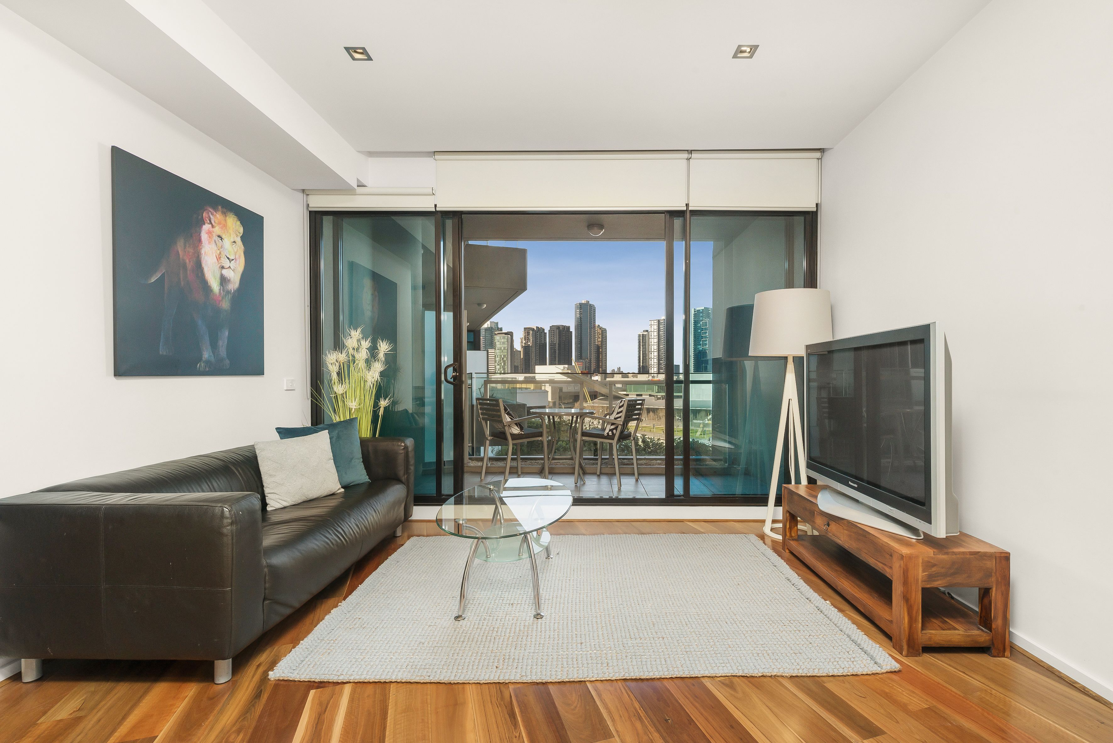 A modern living room with a glass sliding door leading to a balcony, a sofa, a glass coffee table, a TV on a wooden stand, and a cityscape view.