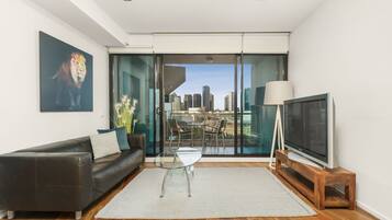 A modern living room with a glass sliding door leading to a balcony, a sofa, a glass coffee table, a TV on a wooden stand, and a cityscape view.