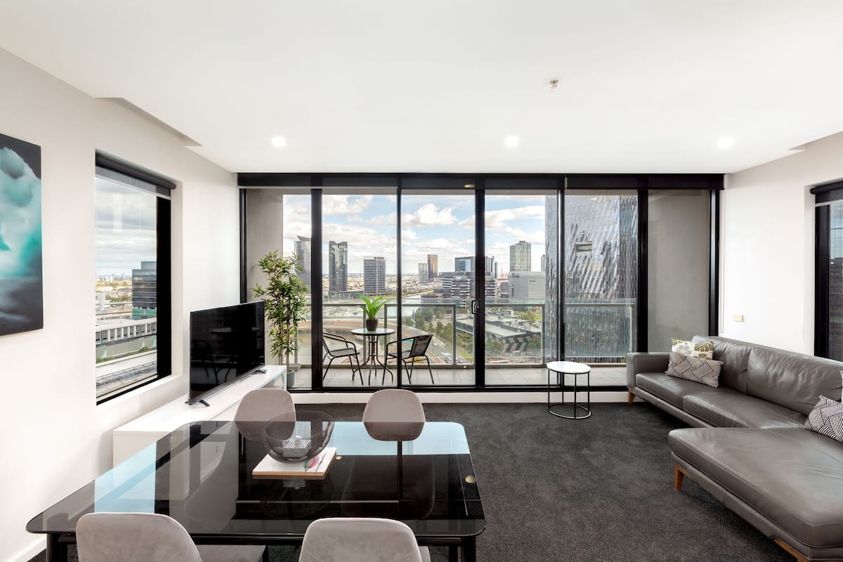 Ein modernes Wohnzimmer mit Glastisch, Sofa und Fernseher. Ein Balkon bietet Blick auf die Stadt.