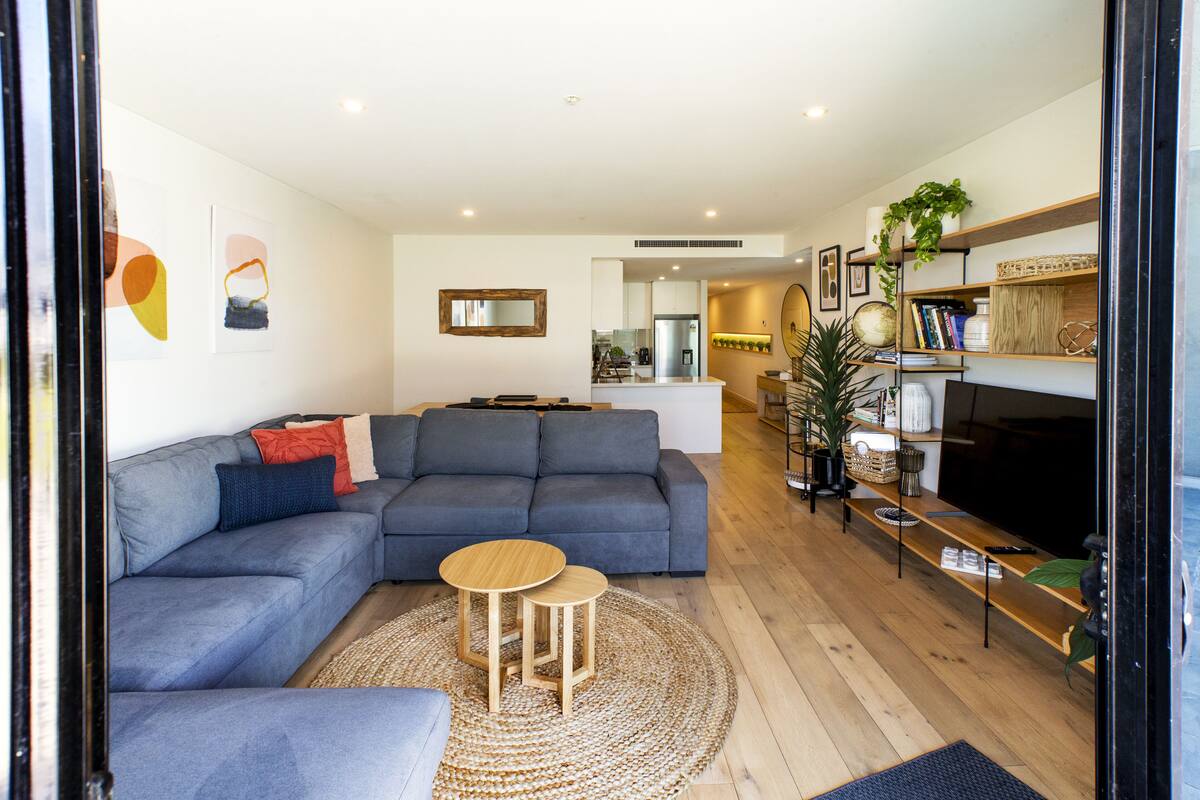 Ein modernes Wohnzimmer mit einem grauen Ecksofa, einem kleinen Holztisch und einer Küchenzeile im Hintergrund.