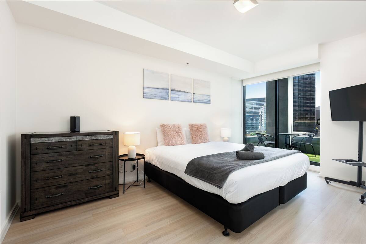 Ein modernes Schlafzimmer mit einem großen Bett, einer Kommode, einem Fernseher und einem Balkon mit Blick auf die Stadt.