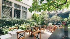 Un área de comedor al aire libre con mesas y sillas, rodeada de follaje y un edificio con ventanas grandes.