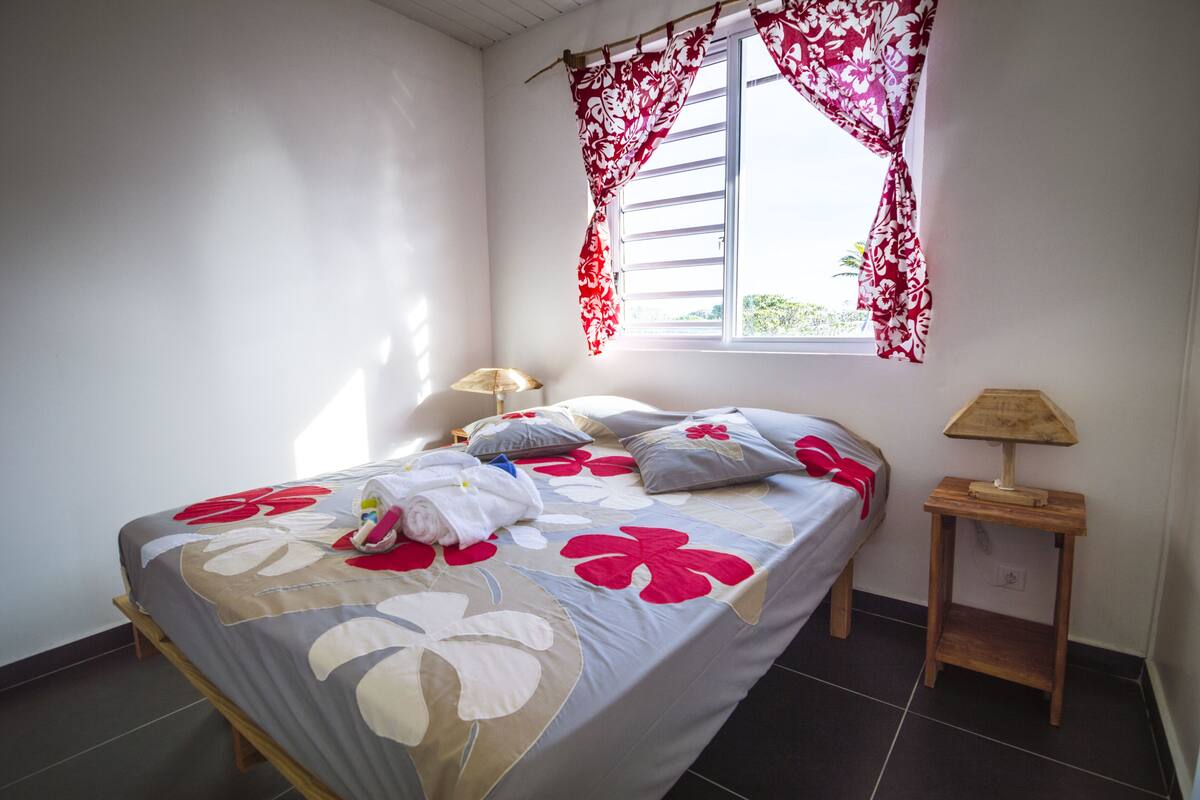 A neatly made bed with floral bedding, a bedside lamp, and a window with red curtains.