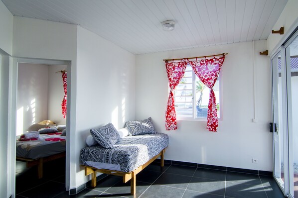 A bedroom with a bed, a window with red curtains, and a door to the right. - Va'a i te Moana (Rangiroa)
