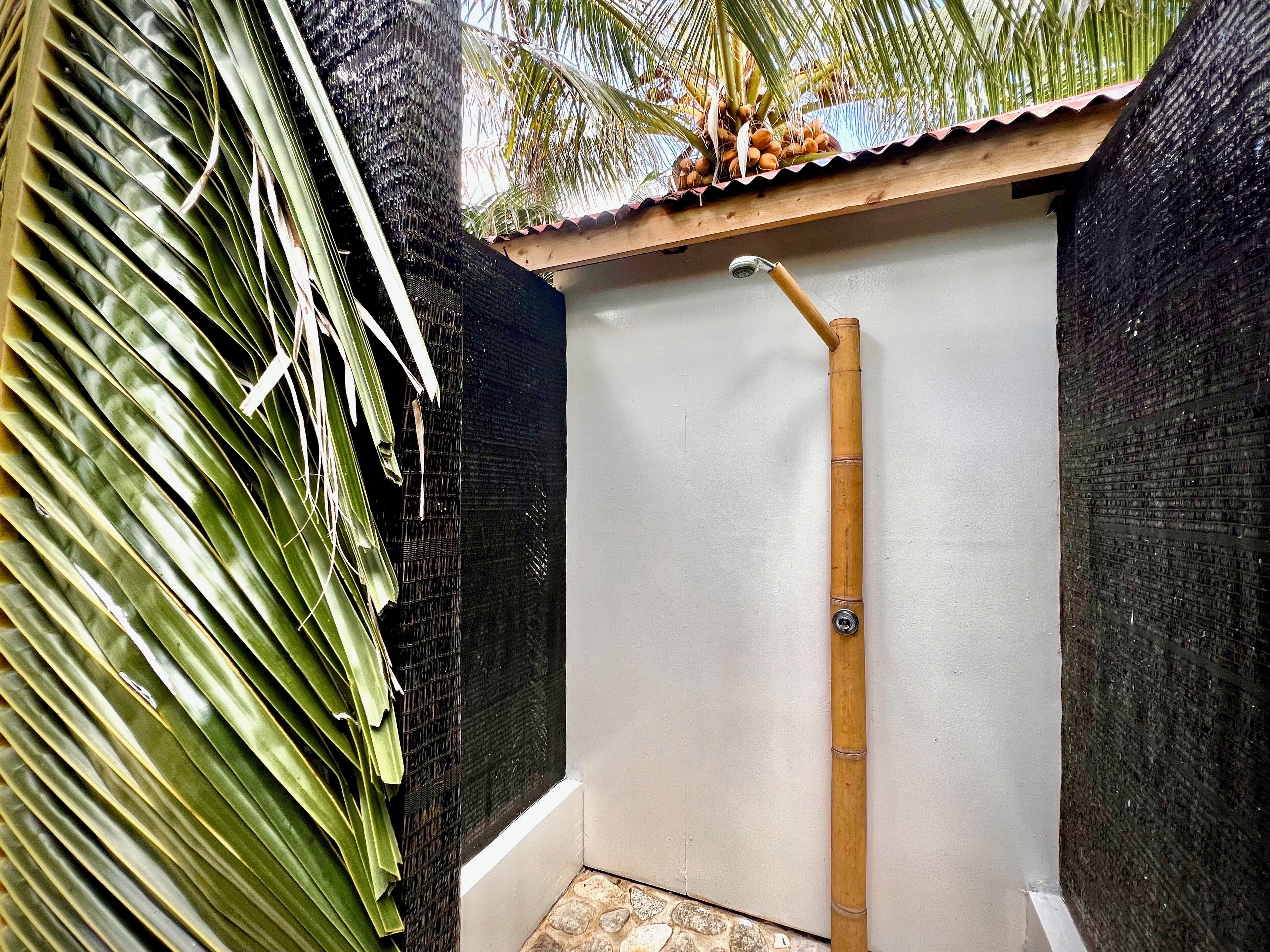 A small, enclosed shower area with a bamboo showerhead, surrounded by dark walls and a palm leaf.