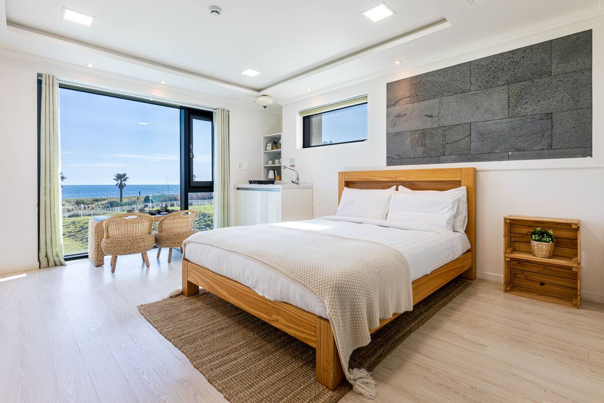 A bedroom with a large bed, a wooden nightstand, a wicker chair, and a view of the beach through a large window.