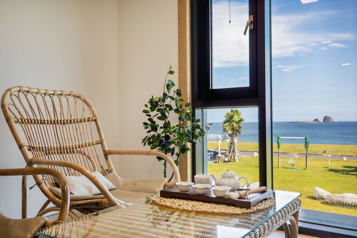 A wicker chair, a table with a tray of tea sets, and a view of the sea through a large window.