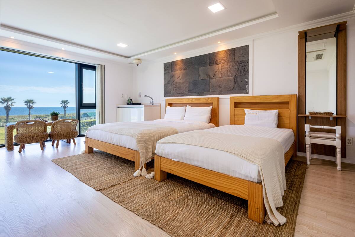 A modern bedroom with two beds, a large window with a view of the beach, and a small kitchenette.