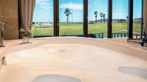 A bathtub with a view of a grassy area and palm trees through large windows. - Sun & Moon Resort (Seogwipo)
