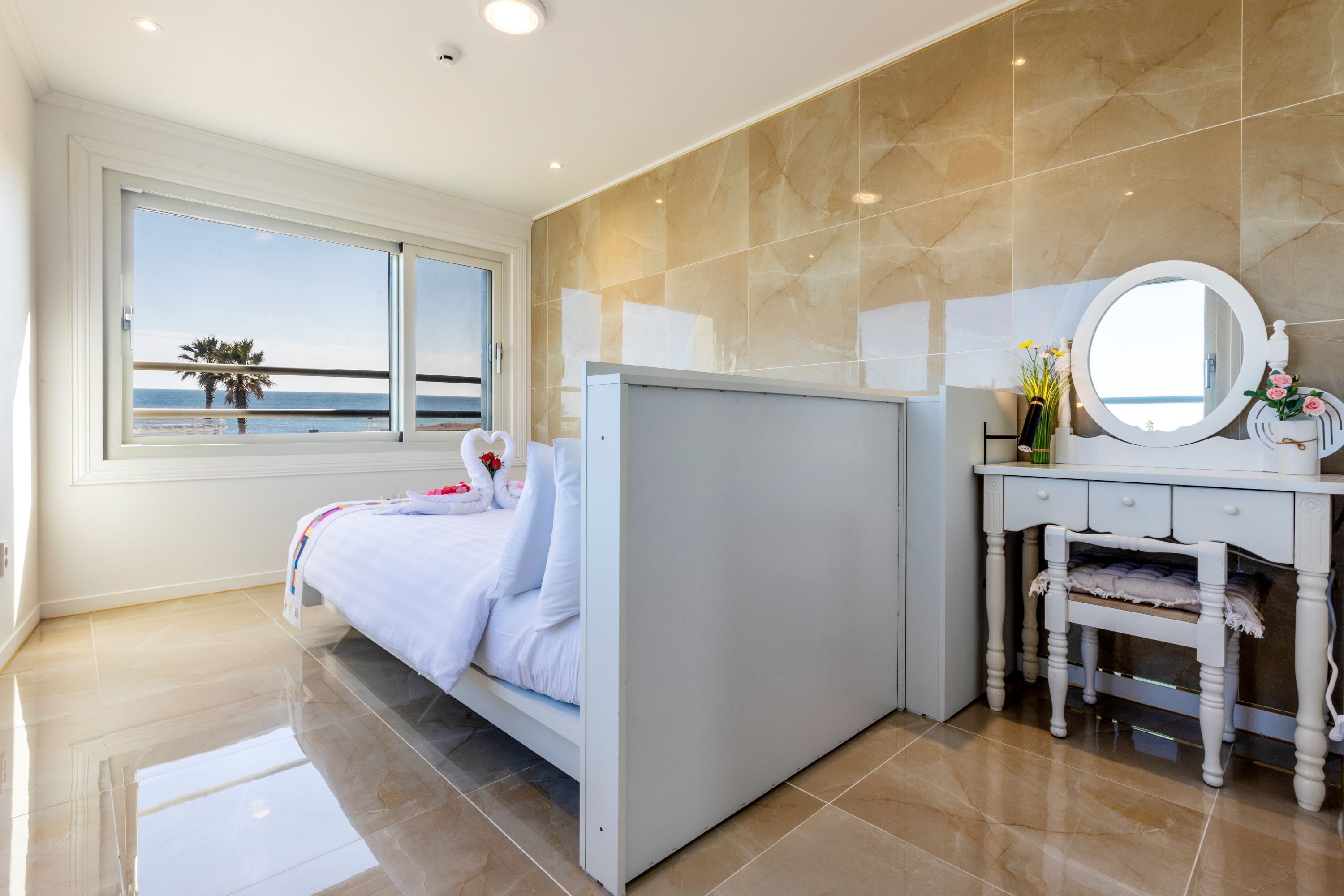 A bedroom with a bed, a vanity with a mirror, and a window with a view of the beach.