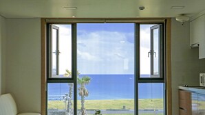 A room with a large sliding glass door leading to a view of the ocean. There is a bed, a small table with a plant, and a kitchen area with cabinets. - Sun & Moon Resort (Seogwipo)