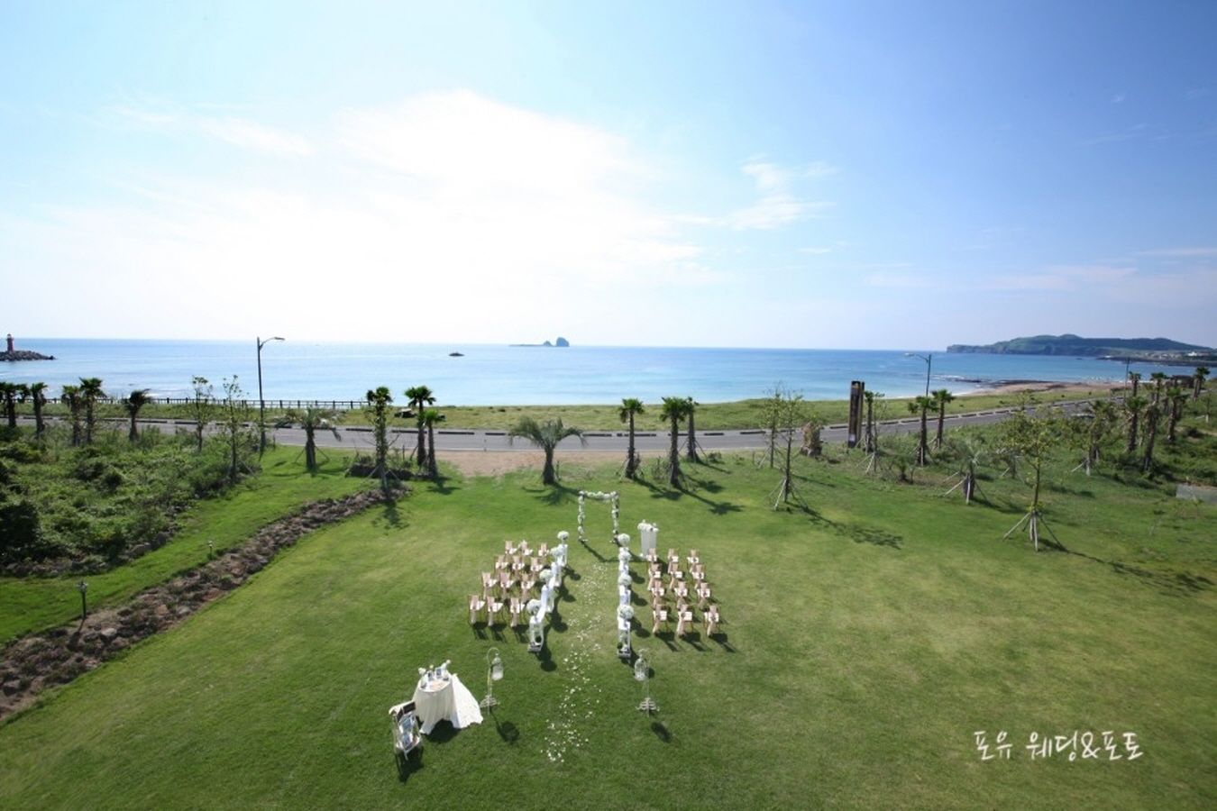 An aerial view of a grassy area with a white tent and tables set up, overlooking a beach and ocean.