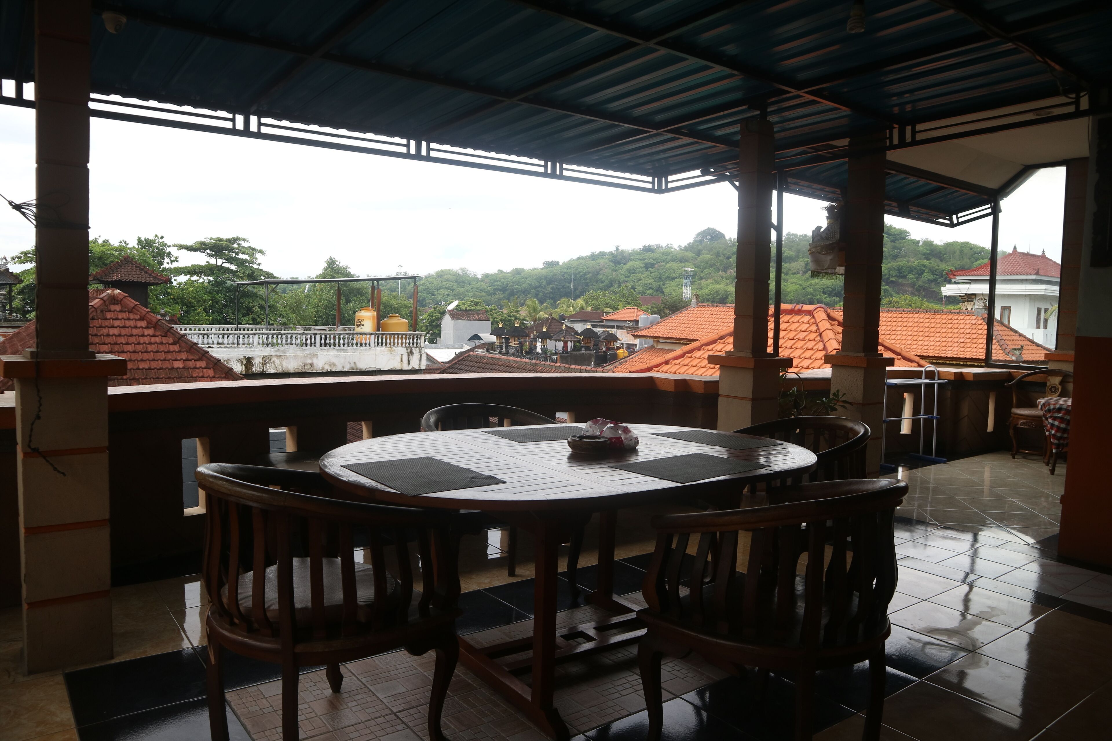 A covered outdoor dining area with a round table and chairs, a tiled floor, and a view of buildings and trees in the background.