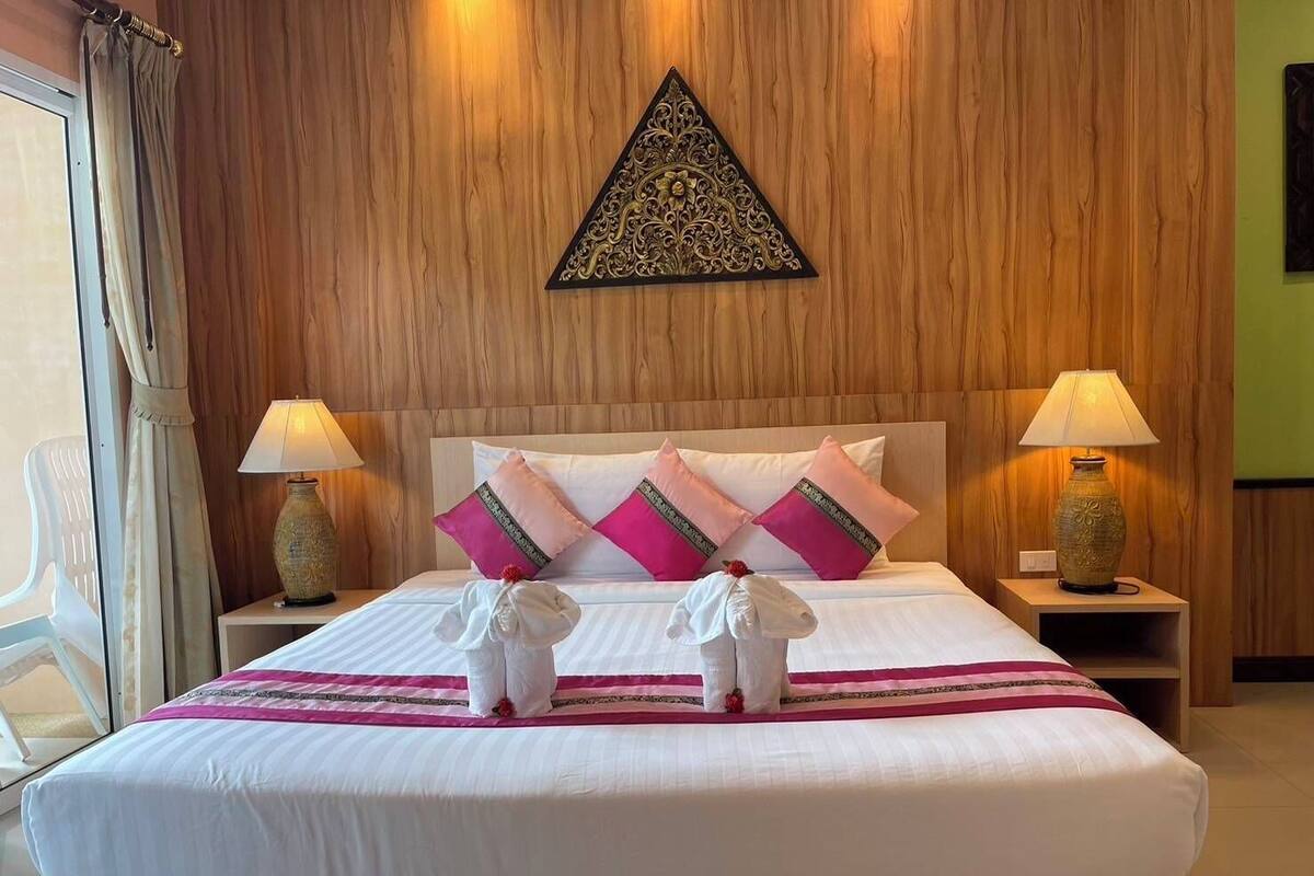 A hotel room with a wooden paneled wall, a bed with white bedding and pink and gold pillows, two bedside lamps, and a decorative wall piece above the bed.