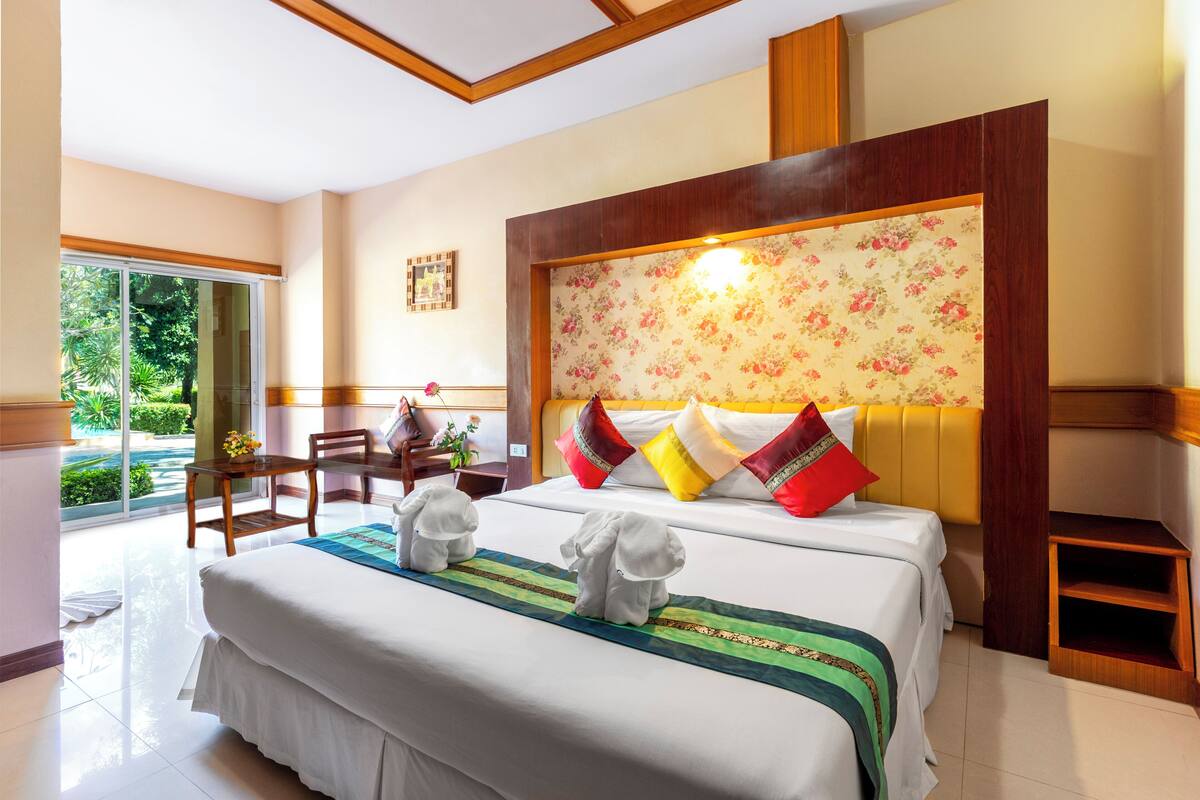 A hotel room with a large bed, a wooden headboard, a dining area with a table and chairs, and a view of the outdoors through a sliding glass door.