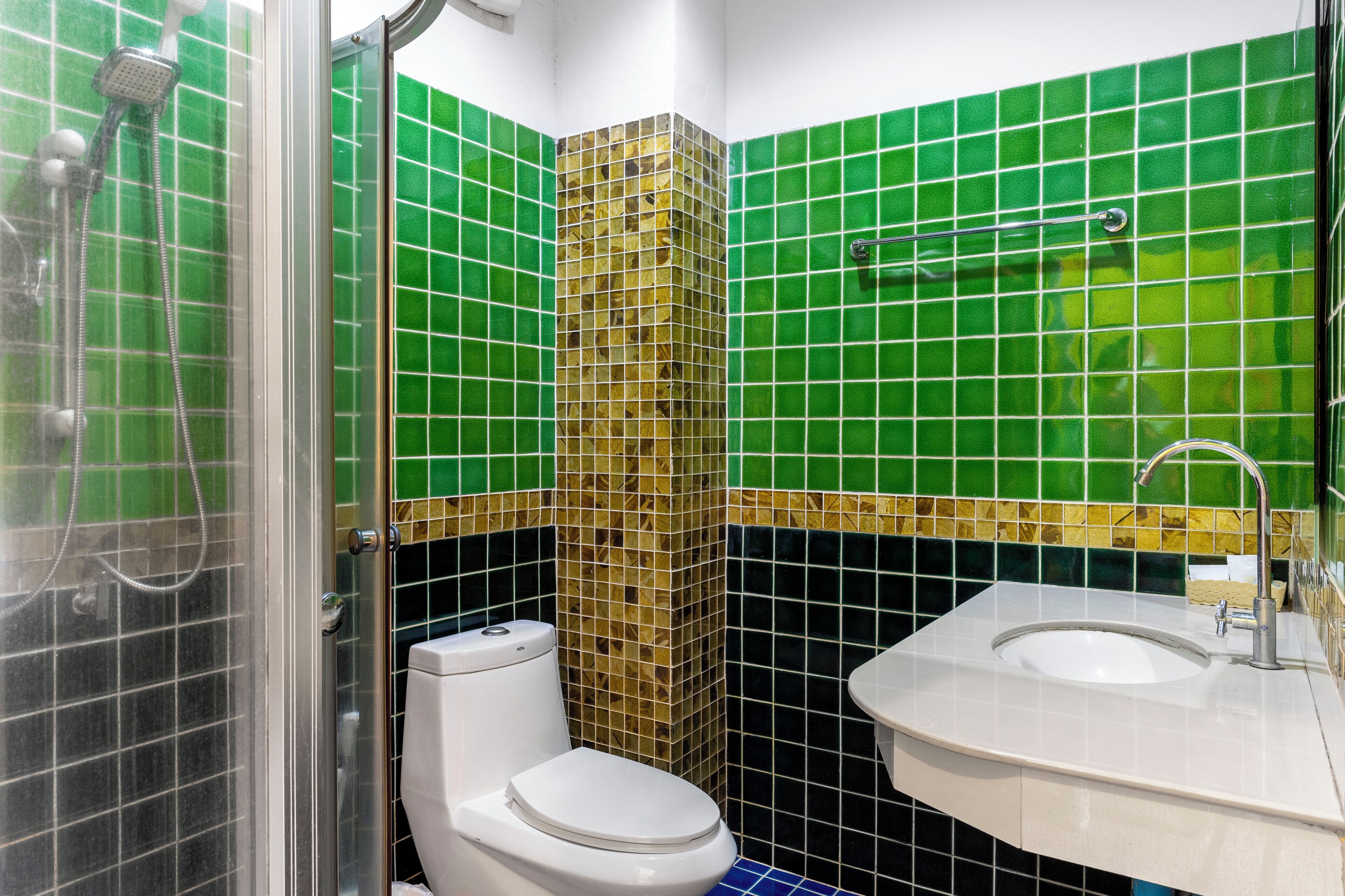 Et badeværelse med vægge belagt med grønne og sorte fliser, et hvidt toilet, en bruseniche og en vask med spejl.